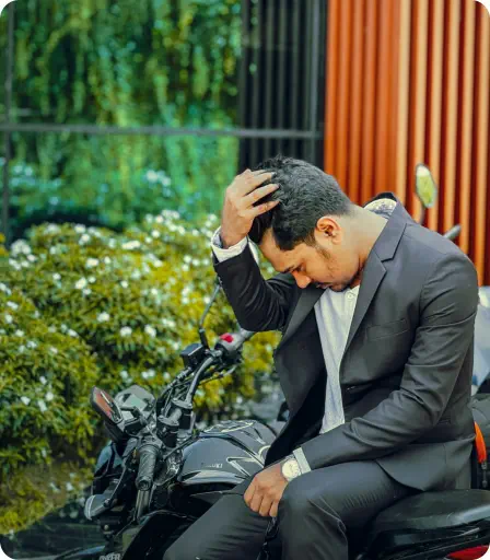 A man in a suit leans on a motorcycle in front of an expensive looking house. He is looking down and holding his head in concern
