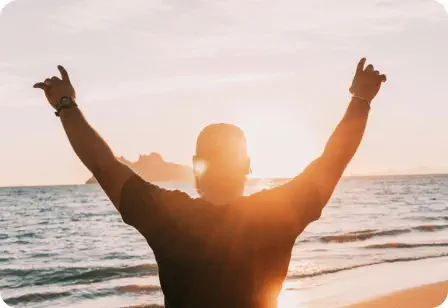 A man faces away watching the sunrise with both hands up in a victory stance