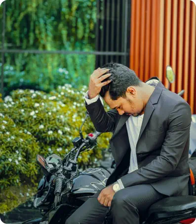 A man in a suit leans on a motorcycle in front of an expensive looking house. He is looking down and holding his head in concern